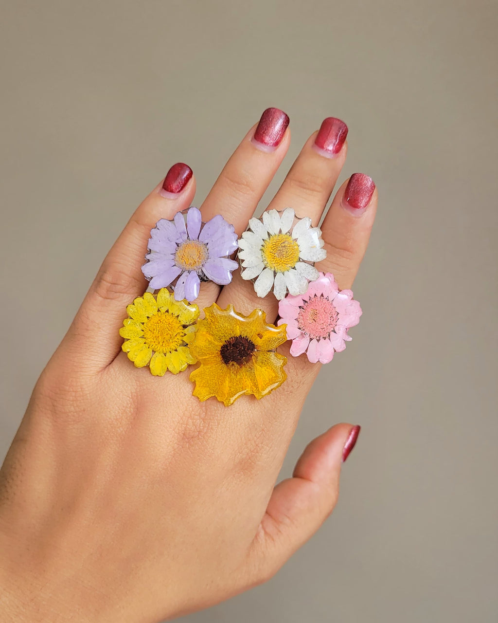 Collection of five pressed-flower resin rings — lilac, white daisy, yellow, sunflower and pink — worn on a hand