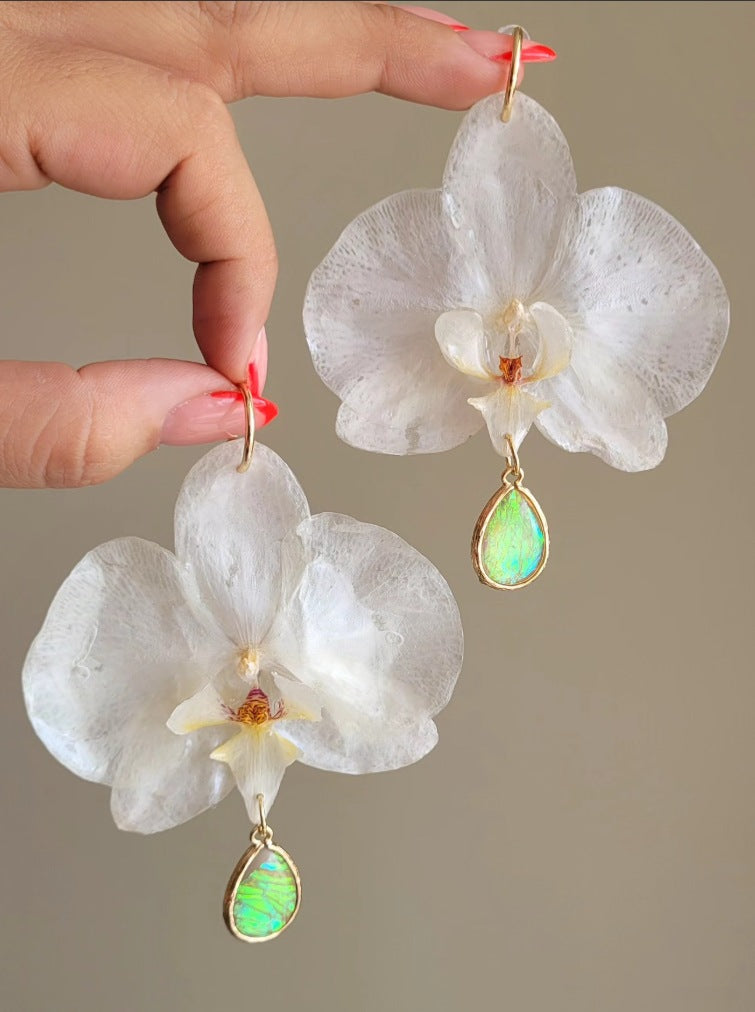 Hands holding a pair of 3D-encapsulated white orchid earrings with green opal teardrops.