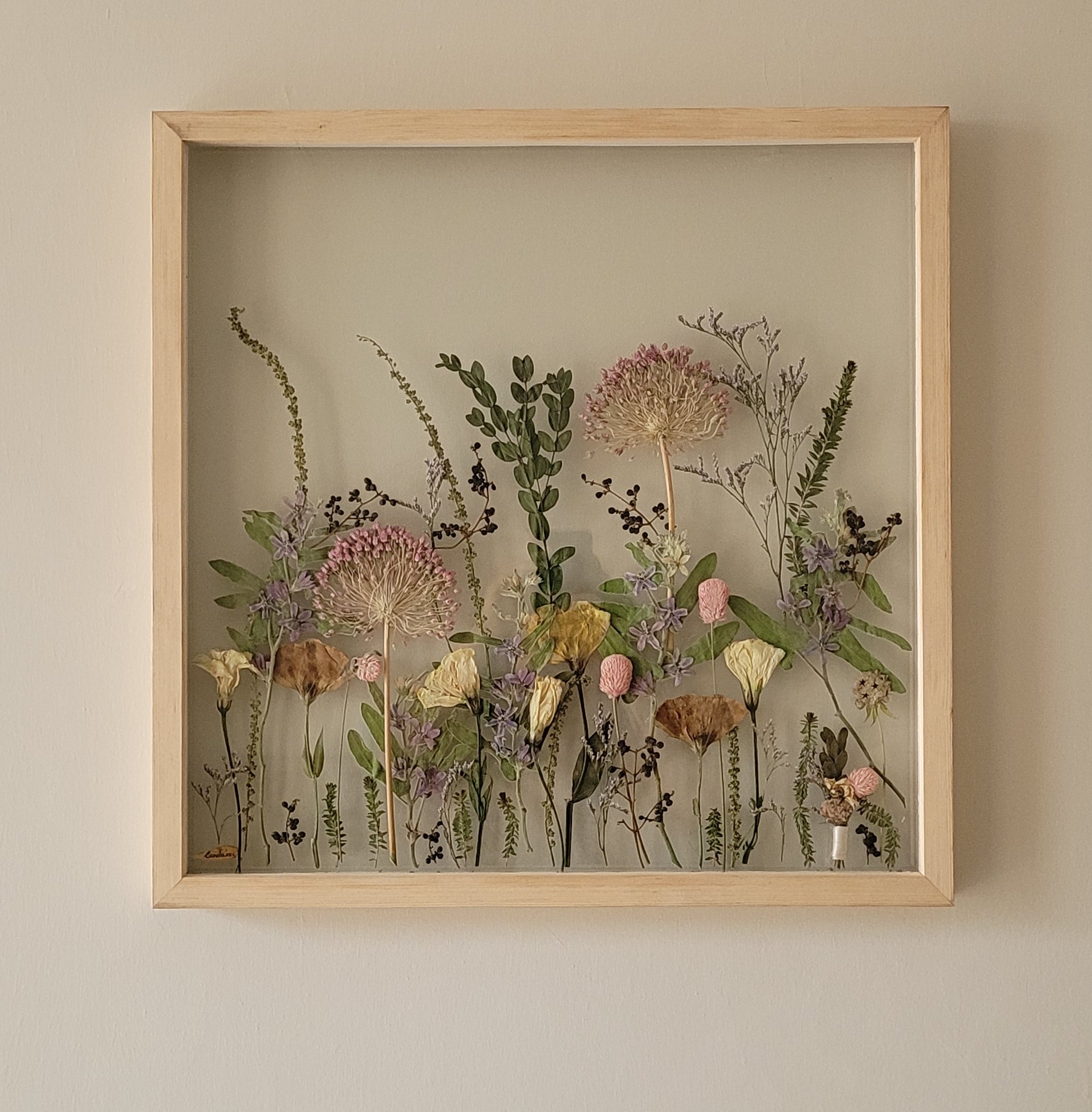 A framed pressed-flower composition arranged like a meadow, hung on a cream wall.