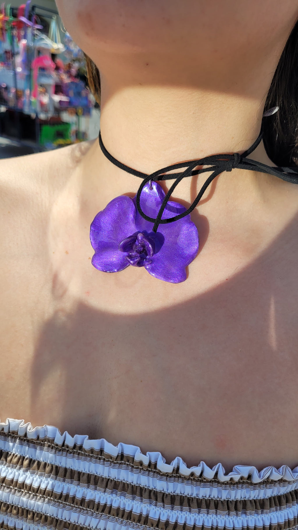A close-up lifestyle shot of a person wearing a black cord choker necklace with a vibrant purple orchid flower pendant, set against a bright, sunlit outdoor background.
