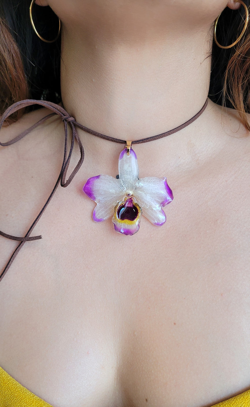 A close-up of a person wearing a brown cord choker necklace with a dangling orchid flower pendant. The flower has white petals with purple edges and a dark purple center with a small gold accent.